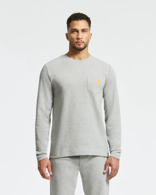 A man is wearing a light gray long-sleeve shirt and matching pants. The shirt, a Psycho Bunny "MENS VAIL WAFFLE LOUNGE LONG SLEEVE TEE - B6T672E200," features a small yellow logo on the chest pocket. The background is plain white.