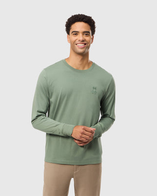 A person wearing the Psycho Bunny MENS ALBERTON LONG SLEEVE TEE - B6T867D200 in sage green, featuring a small embroidered design on the chest, stands against a simple background. They have short curly hair and are smiling with hands clasped in front, creating a fashion masterpiece.