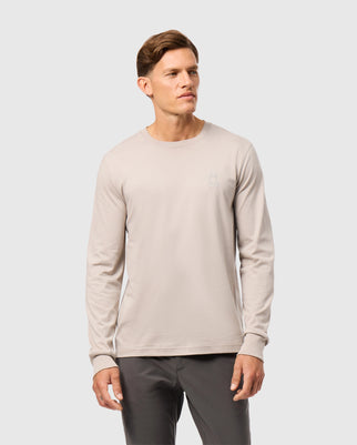 A man wearing a light beige long sleeve tee stands against a plain light gray background. The tee, crafted from Pima cotton and featuring a small embroidered design on the left chest, is from Psycho Bunny's MENS ALBERTON LONG SLEEVE TEE - B6T867D200 collection. He has short brown hair and a neutral expression.