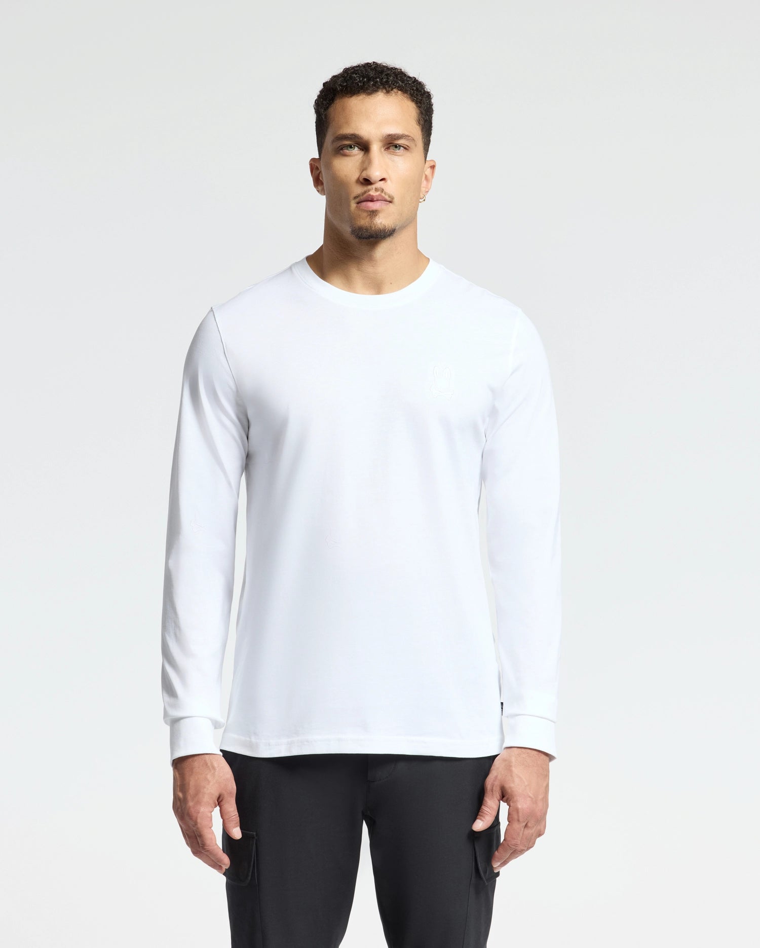 A person stands against a plain background, wearing the Psycho Bunny MENS ALBERTON LONG SLEEVE TEE in Pima cotton and white, paired with black pants. The individual has short curly hair and a calm expression.