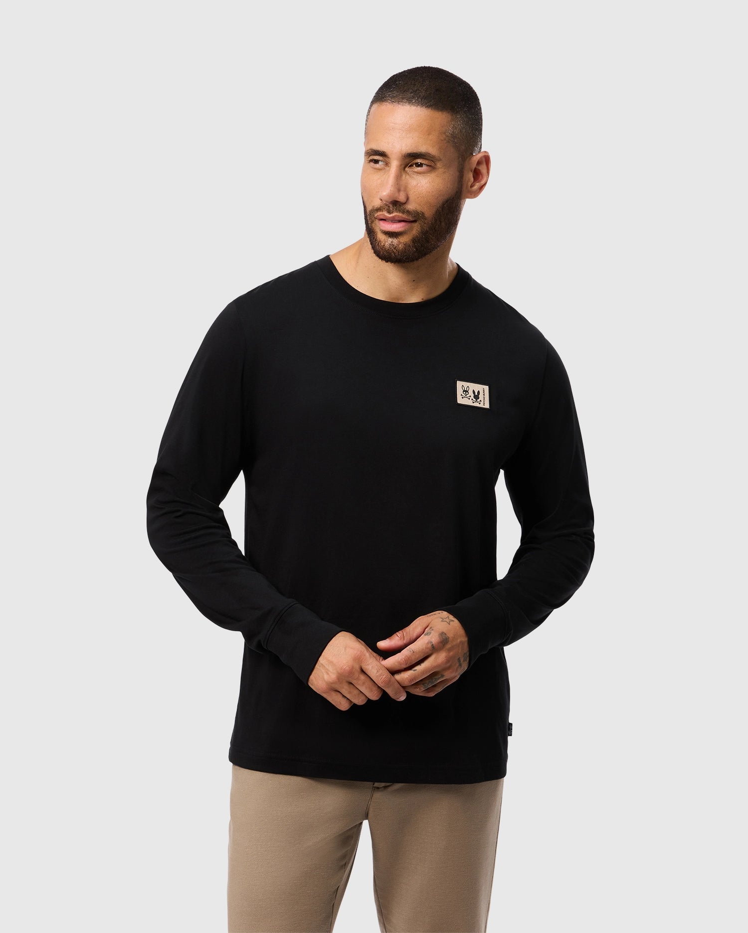 A man with short hair and a beard is wearing the Psycho Bunny Men's Monroe Long Sleeve Tee in black, made of lightweight pima cotton, featuring a small patch on the chest. He pairs it with light brown pants as he stands against a plain white background, looking to the side with a relaxed expression.