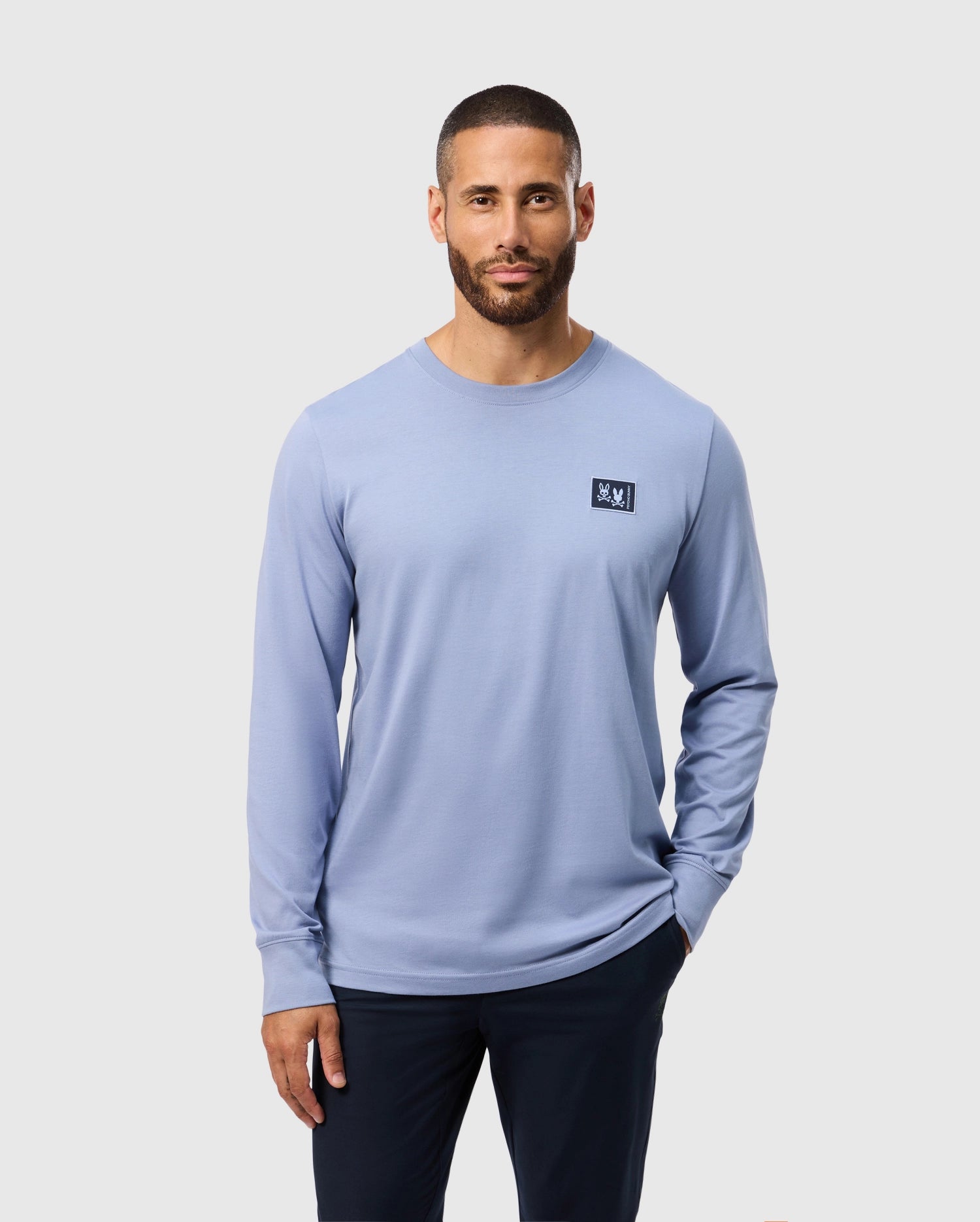 A man with short hair and a beard is wearing the Psycho Bunny MENS MONROE LONG SLEEVE TEE - B6T975D200 in light blue, made from pima cotton. The shirt features a small square logo on the chest. He stands with one hand in his pocket against a plain background, showcasing classic comfort and versatile style.