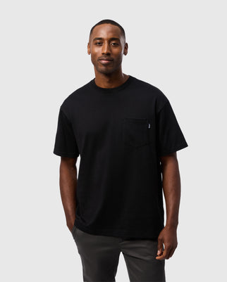 A person stands against a plain, light-colored background wearing a Psycho Bunny MENS CLYDE OVERSIZED POCKET TEE - B6U012D200 in black, made from heavyweight cotton, and dark gray pants. Their hands are casually in their pockets, and they are looking toward the camera.