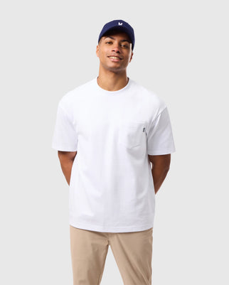 A man stands smiling with his hands behind his back, wearing Psycho Bunny's white MENS CLYDE OVERSIZED POCKET TEE (B6U012D200) made from breathable Peruvian Pima cotton, beige pants, and a navy blue baseball cap. The background is plain light gray.