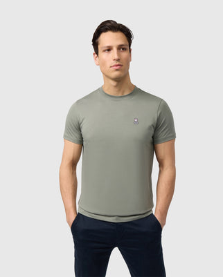 A man standing confidently, wears a Psycho Bunny Men's Classic Crew Neck Tee in solid olive green with a small logo on the left chest, against a plain light background.