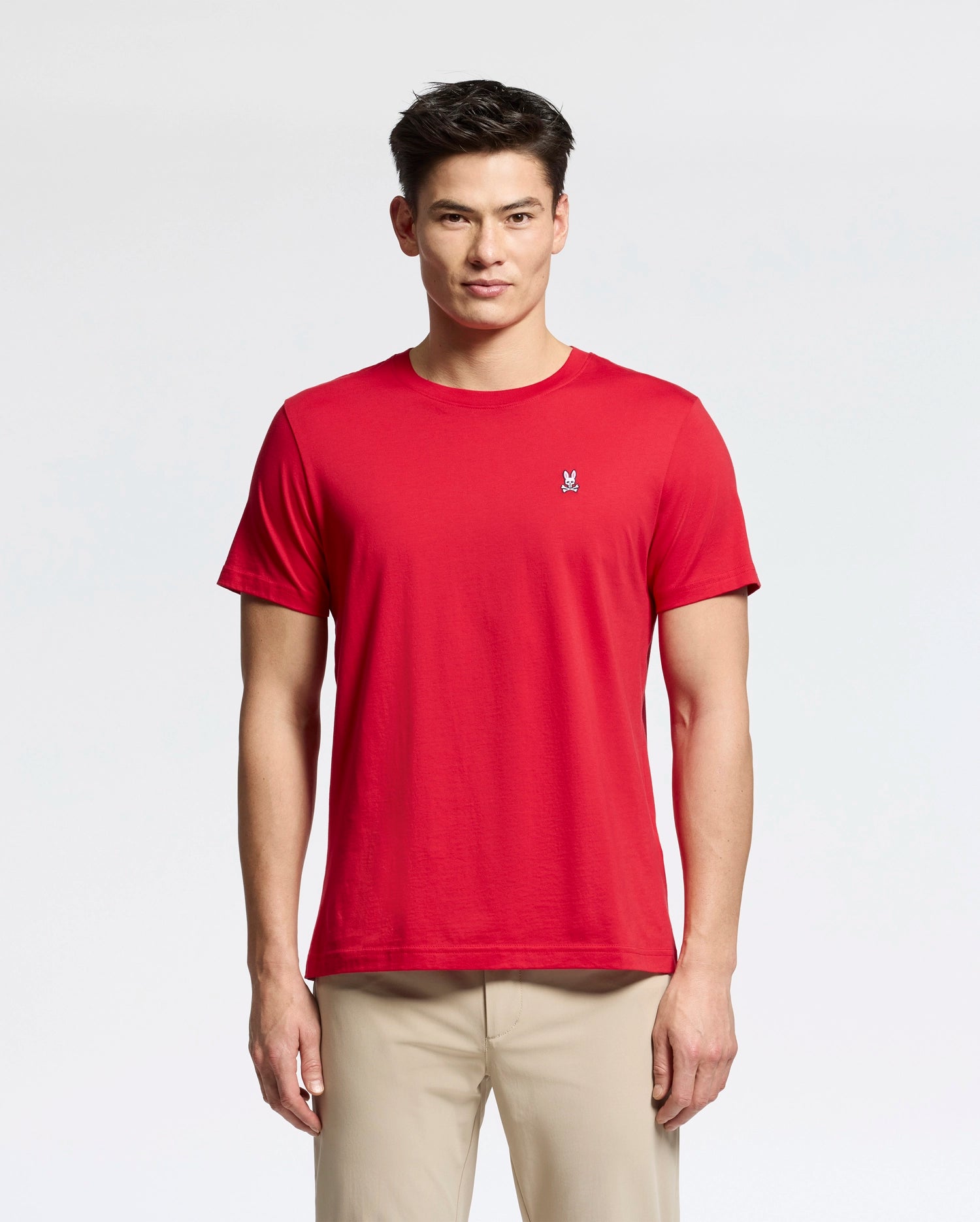 A person in the Psycho Bunny MENS CLASSIC CREW NECK TEE - B6U014CRPC, red with the logo on the left chest, stands against a white background wearing light beige pants and has short, styled hair.