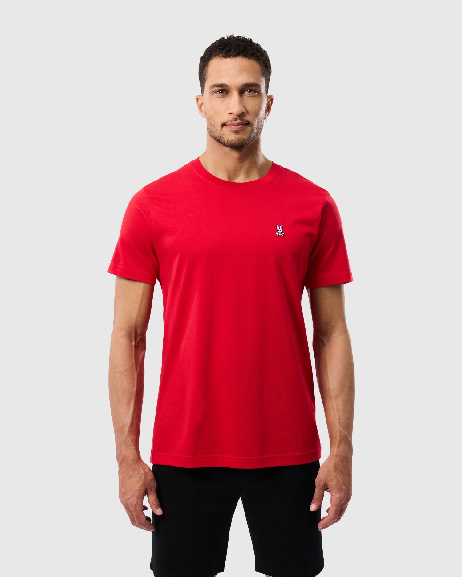 A man with short dark hair and a short beard is wearing a fitted red Psycho Bunny MENS CLASSIC CREW NECK TEE - B6U014CRPC made from 100% Pima cotton, featuring a small white logo on the left chest, paired with black shorts. He stands against a plain, light gray background, looking directly at the camera with a neutral expression.