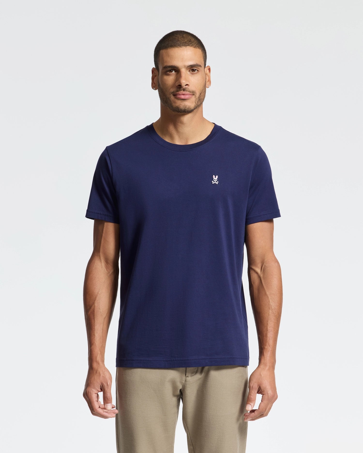 A man wearing the versatile MENS CLASSIC CREW NECK TEE - B6U014CRPC in blue by Psycho Bunny, featuring a small white logo on the chest, stands against a plain white background. He is paired with beige pants and has short hair.