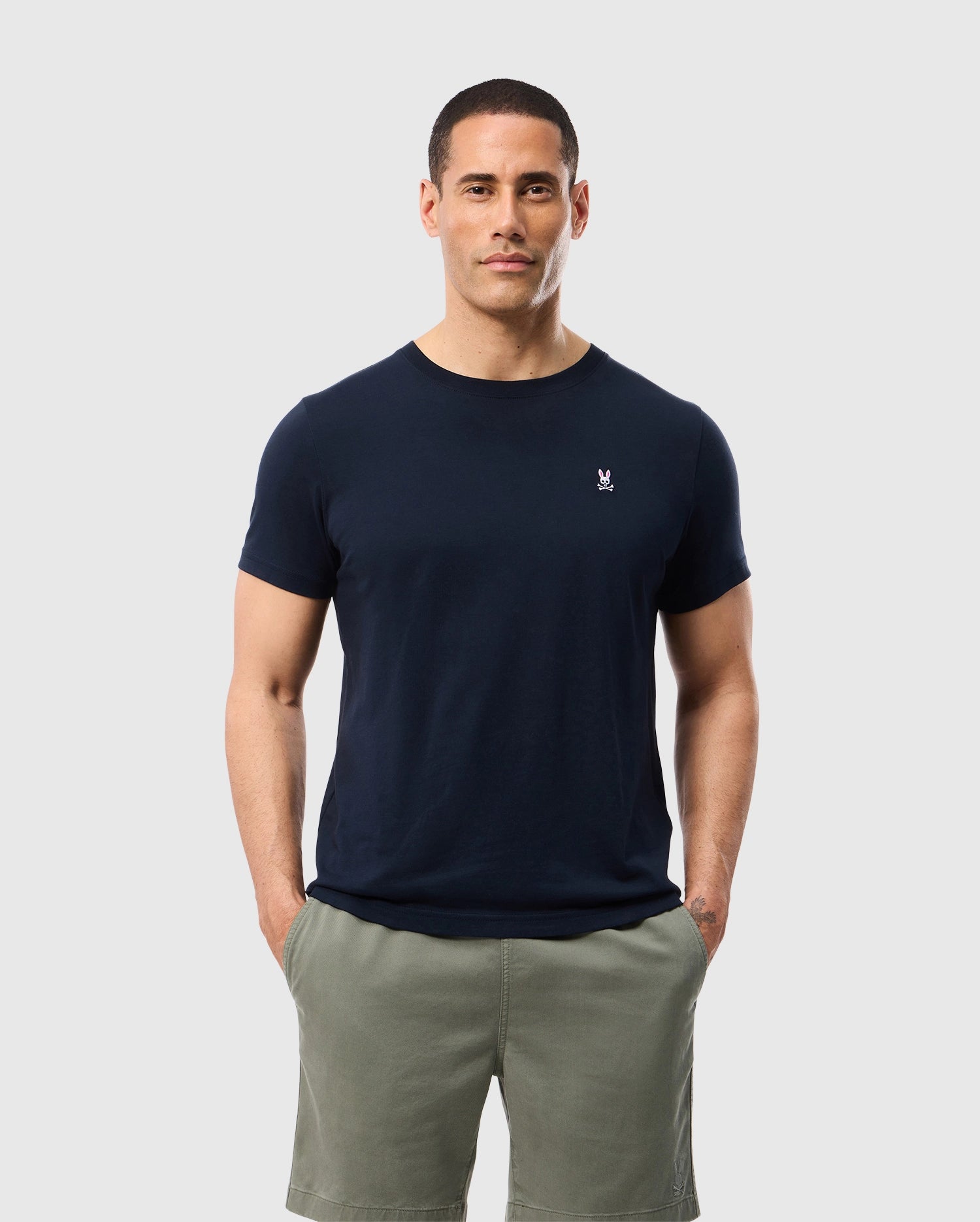 A man with short hair is standing against a plain background, wearing a navy blue Men's Classic Crew Neck Tee (B6U014CRPC) by Psycho Bunny, made of 100% Pima cotton and featuring a small logo on the left chest, paired with olive green shorts. He has his hands in his pockets and is looking directly at the camera with a neutral expression.