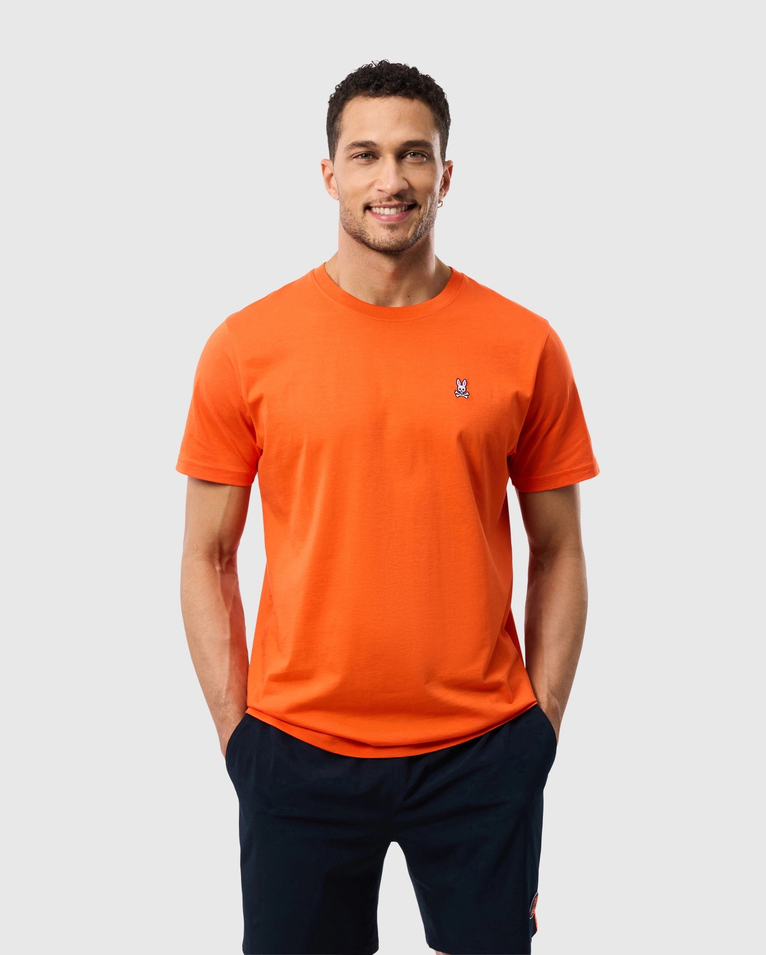 A man with short curly hair is wearing an orange Psycho Bunny Men's Classic Crew Neck Tee (B6U014CRPC), made of Pima cotton and featuring a small embroidered bunny logo on the left side of the chest. He has his hands in the pockets of his dark-colored pants and is smiling at the camera against a plain light gray background.