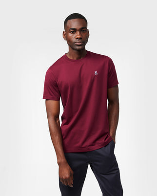 A man stands against a plain white background wearing a maroon Psycho Bunny MENS CLASSIC CREW NECK TEE - B6U014Z1PC crafted from breathable and lightweight Pima cotton, featuring a small logo on the chest and paired with dark pants. He has a neutral expression, with his left hand in his pocket and his right arm resting by his side.