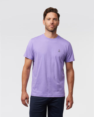 A man wearing the MENS CLASSIC CREW NECK TEE - B6U014Z1PC by Psycho Bunny, featuring a small bunny logo on the left chest, stands against a plain grayish background. He has short brown hair and is wearing dark pants.