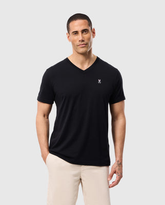 A person is posed against a plain backdrop, wearing the Psycho Bunny MENS CLASSIC V NECK TEE - B6U100ARPC in black, composed of 100% Pima cotton, paired with beige pants. The shirt is adorned with a subtle white Psycho Bunny emblem on the upper left side. The individual has short hair and looks at the camera with a neutral expression.