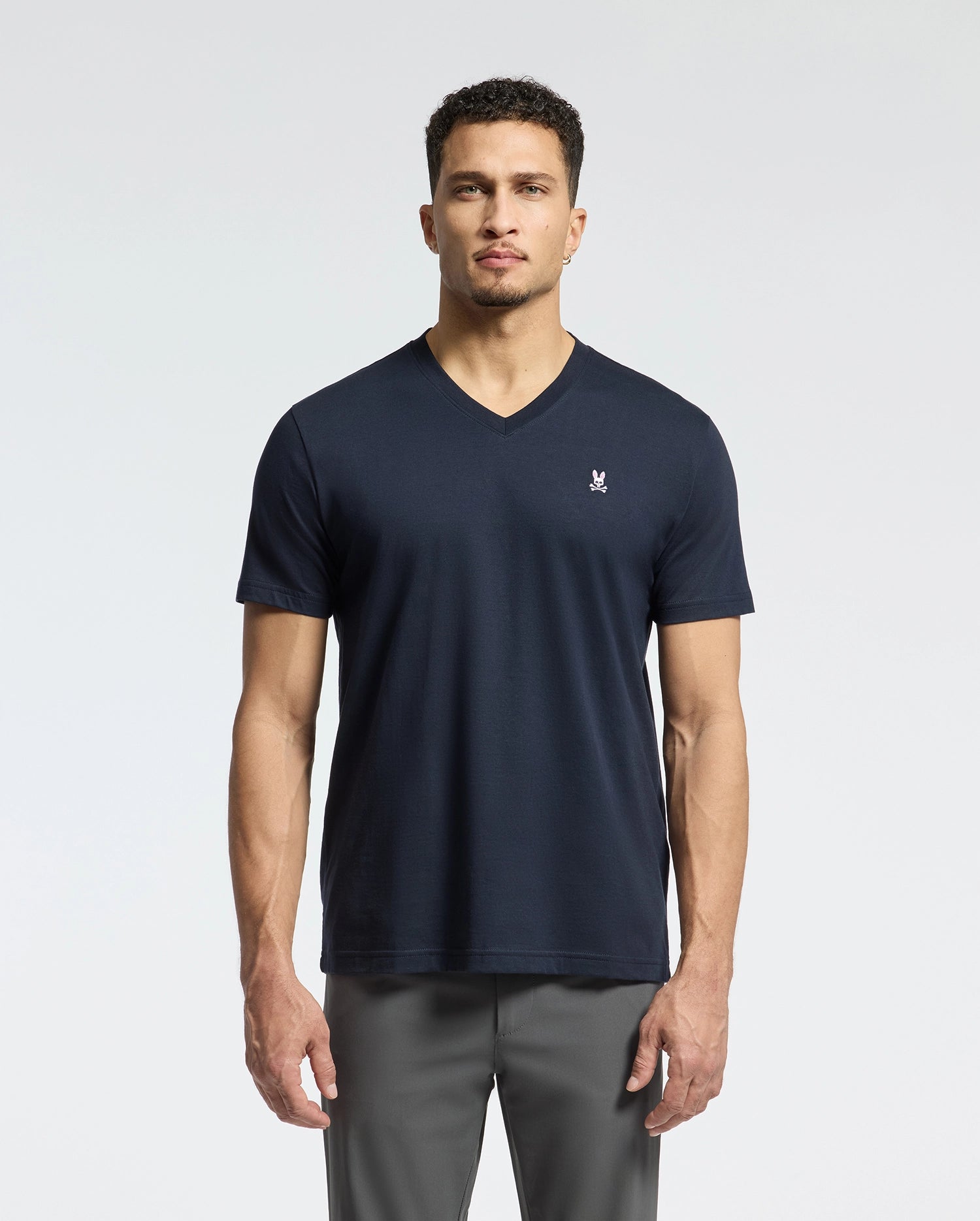 A person is wearing the sleek black MENS CLASSIC V NECK TEE - B6U100ARPC by Psycho Bunny, crafted from soft Pima cotton and adorned with a subtle chest emblem. They're paired with gray pants set against a plain, light-colored backdrop.