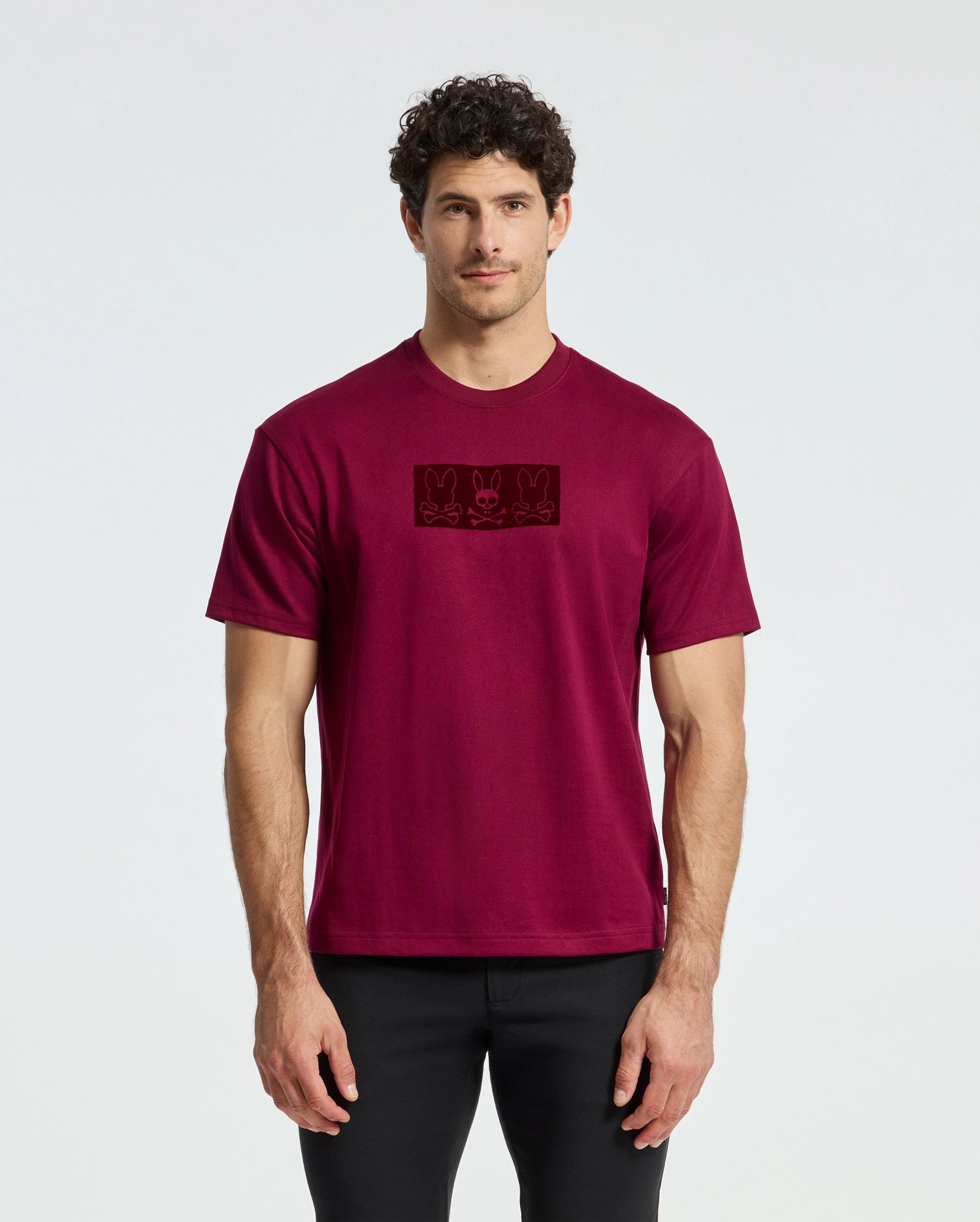 A man with curly hair is wearing a comfortable, relaxed-fit red tee from Psycho Bunny, specifically the MENS ROMEO HEAVY WEIGHT RELAXED FIT TEE (B6U110E200), which features three small designs on the front. He pairs this casual shirt with black pants and stands against a plain white background.