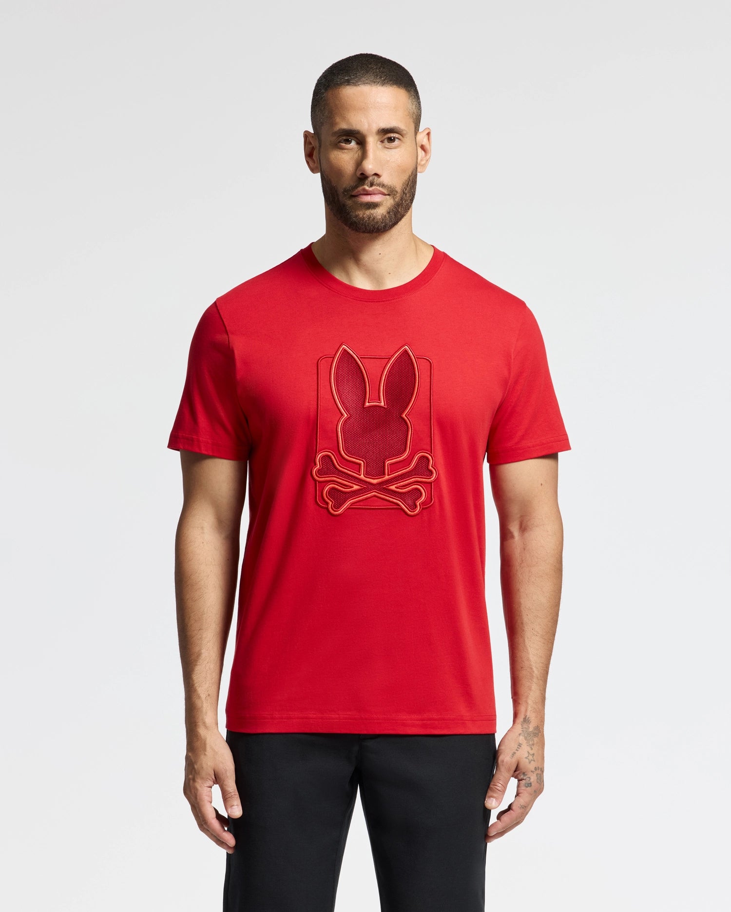 A man with short hair and a beard is wearing a red Psycho Bunny MENS PIERCE EMBROIDERED GRAPHIC TEE - B6U115E200, made from soft Pima cotton. The shirt showcases an embroidered design of a rabbit head above crossed bones. He is standing against a plain white background, paired with black pants.