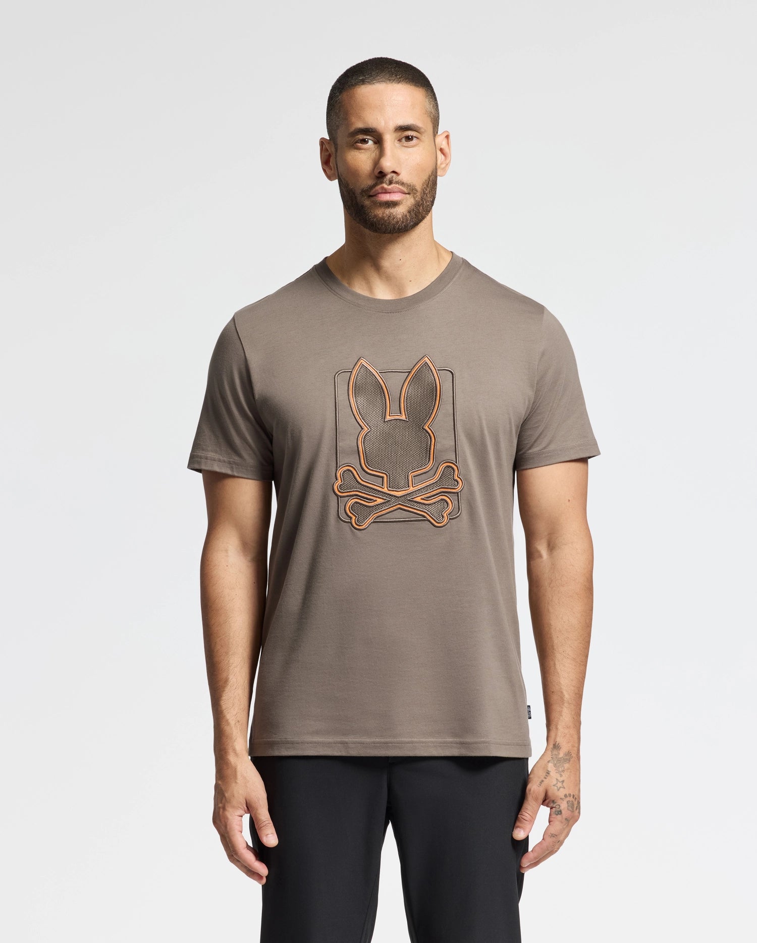 A man with short hair and a beard wears the Psycho Bunny Gray MENS PIERCE EMBROIDERED GRAPHIC TEE - B6U115E200 in soft Pima cotton. It showcases a quirky, orange-outlined rabbit head above crossed bones as part of its unique embroidery, adding a touch of playfulness to his style. He stands against a plain white background, exuding casual confidence.