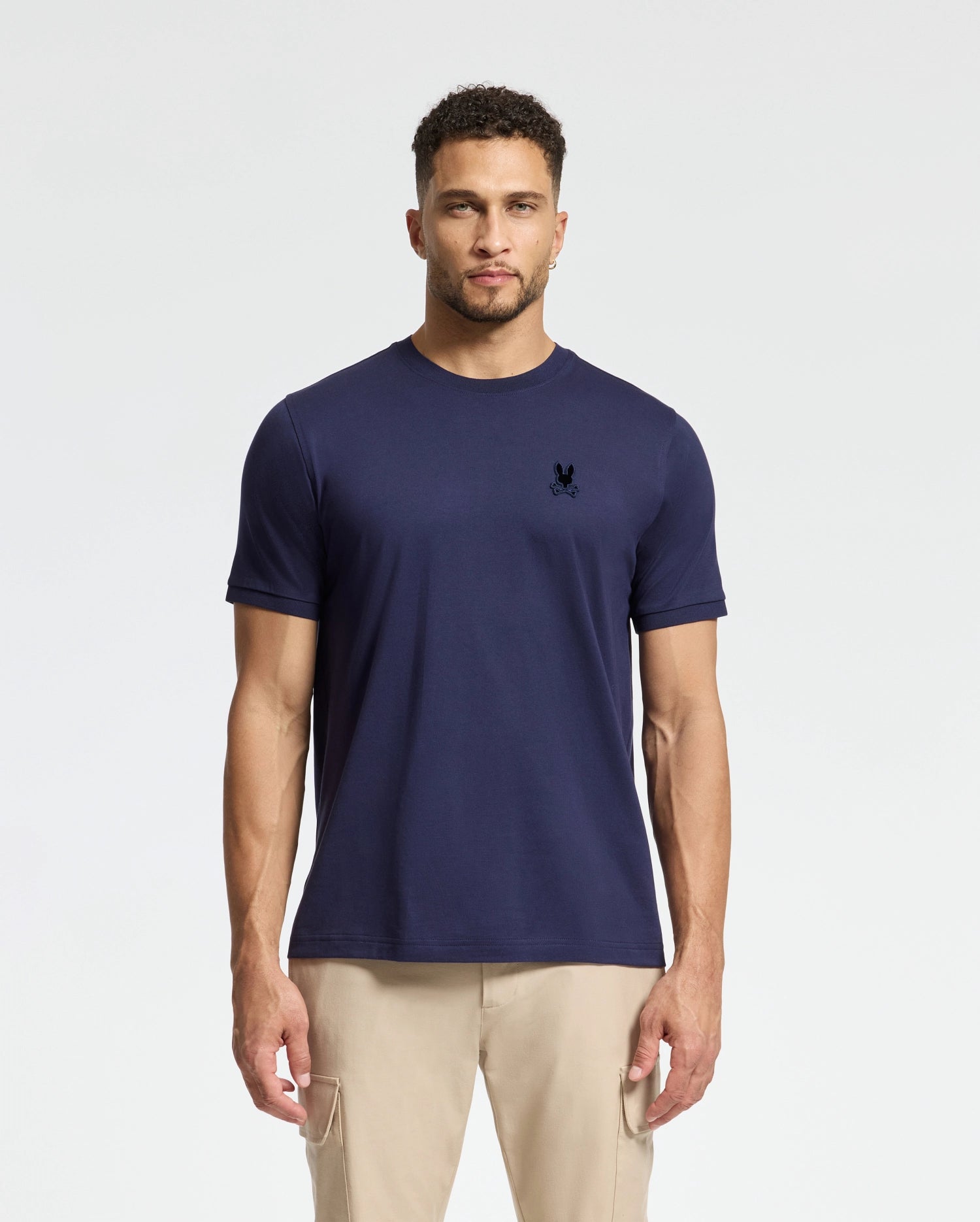 A man in a navy blue Psycho Bunny MENS BRANSON FASHION TEE - B6U137E200 stands facing the camera against a plain background. The tee, crafted from men's Pima cotton, features a small embroidered logo on the left chest. He is wearing beige pants and has short curly hair.