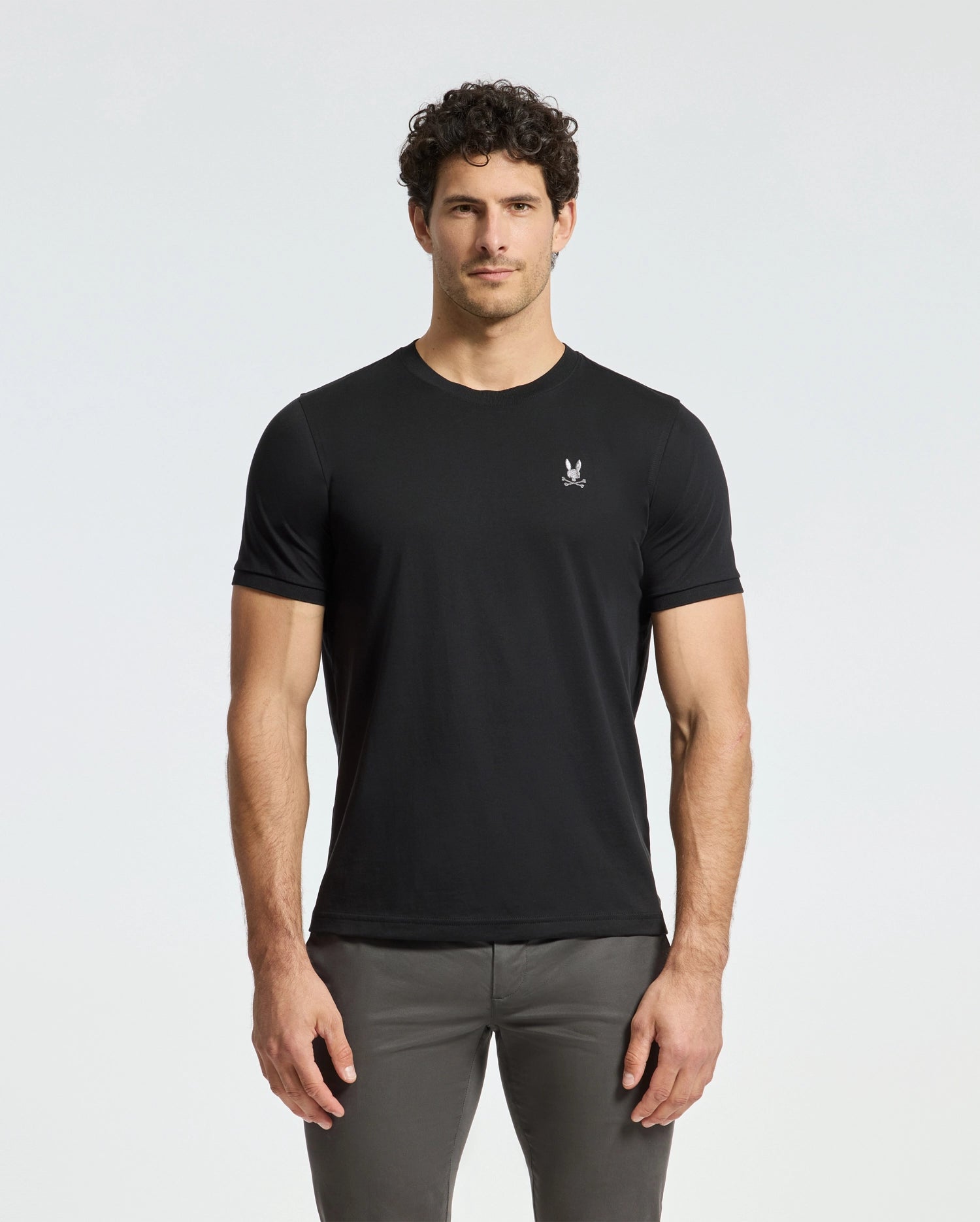 A man with curly hair is wearing a black Psycho Bunny men's Alexander fashion tee, identified by product code B6U139E200, which features a small white logo on the chest, paired with gray pants. He stands against a plain light background, exemplifying luxurious men's fashion with effortless style.