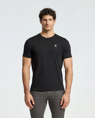 A man with curly hair is wearing a black Psycho Bunny men's Alexander fashion tee, identified by product code B6U139E200, which features a small white logo on the chest, paired with gray pants. He stands against a plain light background, exemplifying luxurious men's fashion with effortless style.