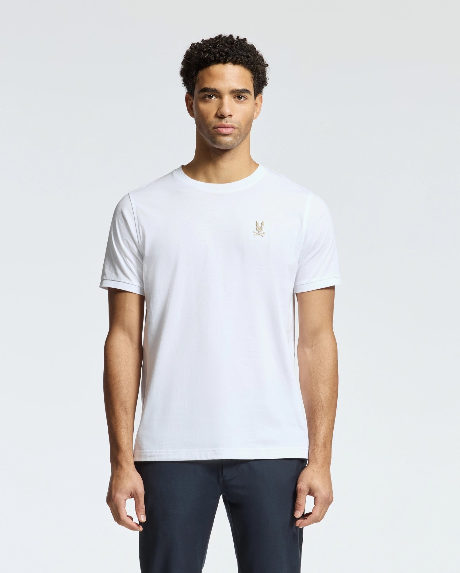 A man with curly hair is sporting the stylish Psycho Bunny MENS ALEXANDER FASHION TEE - B6U139E200, crafted from Pima cotton and adorned with a small embroidered logo on the chest. He pairs it with dark pants, standing against a plain light gray background, radiating effortless style.