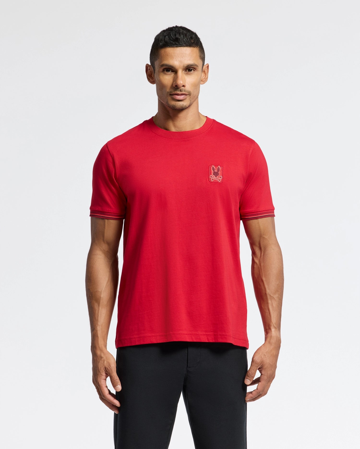 A man stands facing forward wearing the Psycho Bunny MENS PIERCE FASHION TEE - B6U140E200 in red, adorned with a small embroidered bunny patch on the chest. Made from 100% pima cotton, he pairs it with black pants. The neutral background highlights his casual outfit.