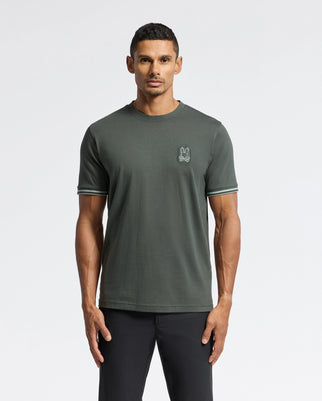 A person stands wearing a dark green MENS PIERCE FASHION TEE - B6U140E200 by Psycho Bunny, featuring a small embroidered logo on the left chest, paired with black pants. This Pima Cotton tee offers a sleek and comfortable fit against the plain light gray background.