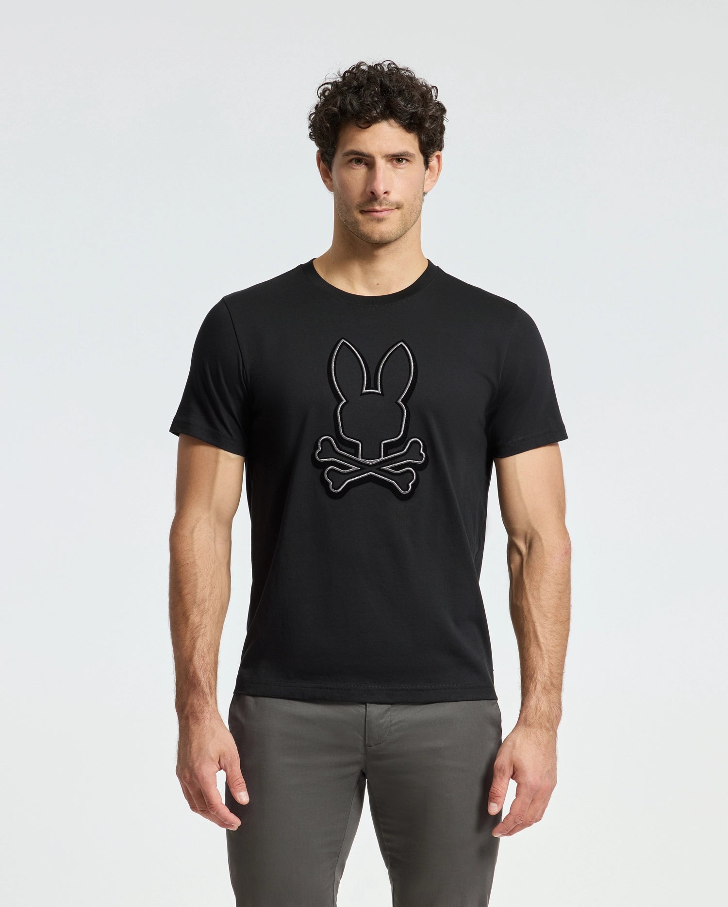 Wearing the stylish Alexander Graphic Tee by Psycho Bunny, this curly-haired man stands out against a plain backdrop. The black tee, made from premium pima cotton, features a striking rabbit head design with crossed bones. Paired with gray pants, his modern and captivating style is effortlessly chic.