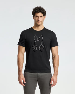 Wearing the stylish Alexander Graphic Tee by Psycho Bunny, this curly-haired man stands out against a plain backdrop. The black tee, made from premium pima cotton, features a striking rabbit head design with crossed bones. Paired with gray pants, his modern and captivating style is effortlessly chic.