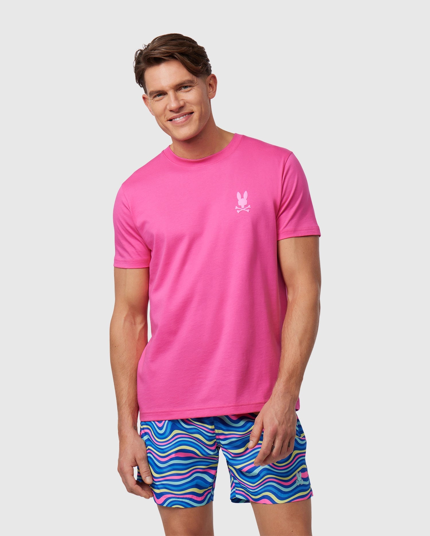 A man smiling, wearing a bright pink Psycho Bunny Pima cotton t-shirt and colorful striped shorts, standing against a plain white background.
