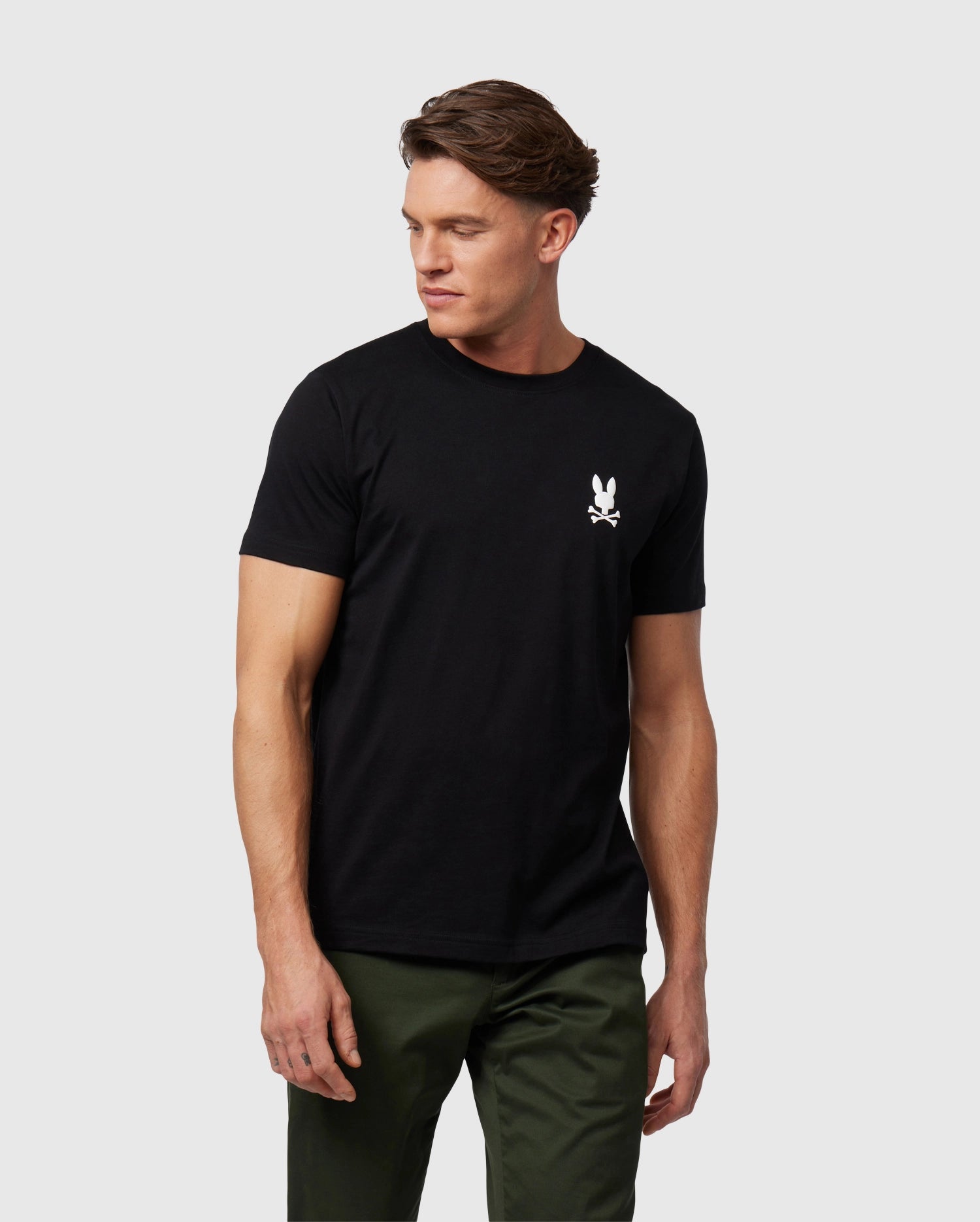 A man standing, wearing a black Psycho Bunny Pima cotton T-shirt with a white logo on the chest and dark green pants, looking to his left with a neutral expression.