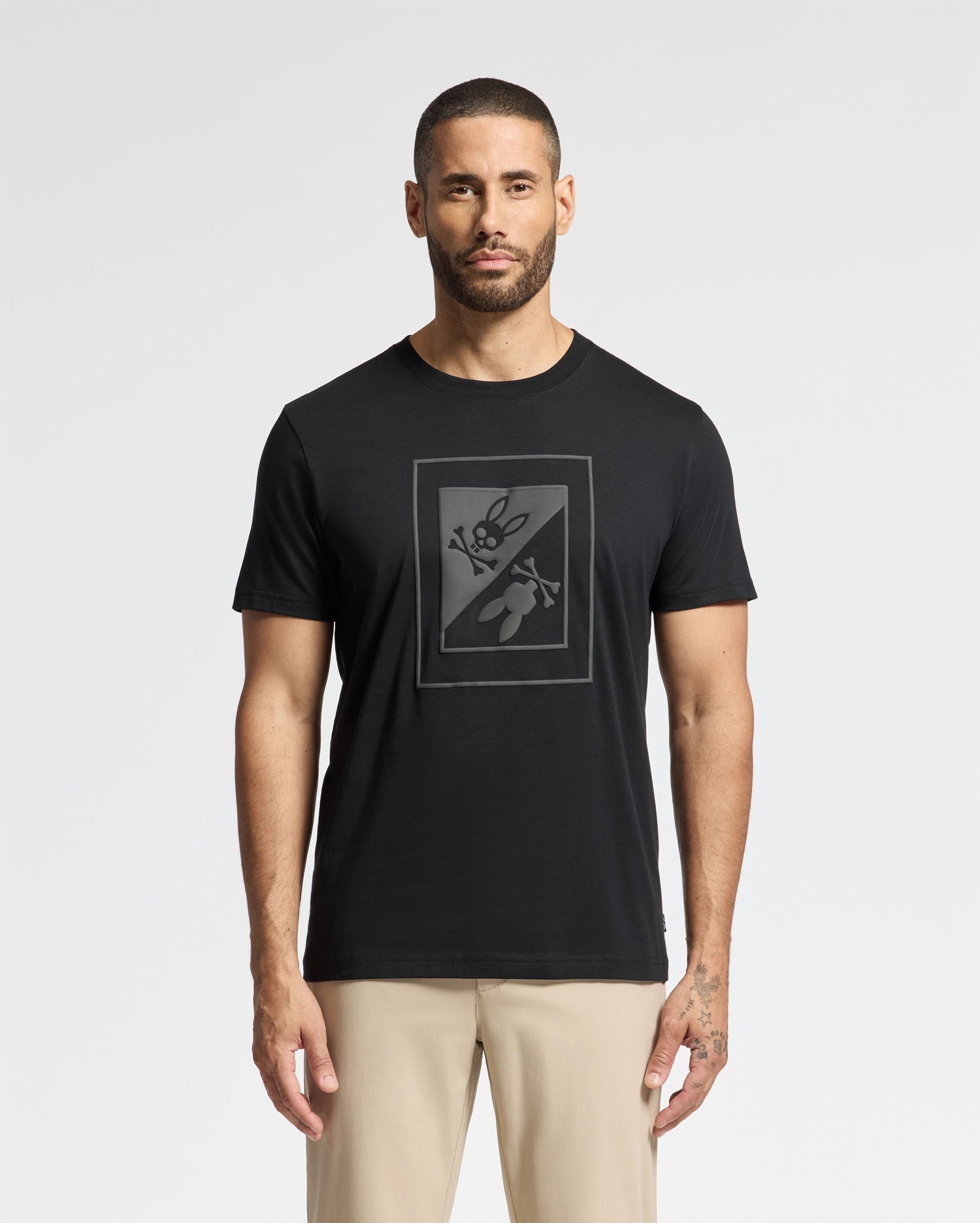 A man with a beard sports the trendy black MENS DOTSERO GRAPHIC TEE (B6U197E200) by Psycho Bunny, showcasing a rectangular graphic of crossed scissors and a bee. He pairs it with beige pants, exuding both comfort and style against a plain white background.