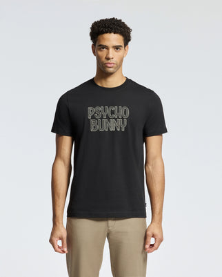 Man in a black "Psycho Bunny" MENS MONARC WORDMARK GRAPHIC TEE - B6U210E200, made from 100% Pima cotton and designed for a regular fit, paired with beige pants, standing against a neutral background.