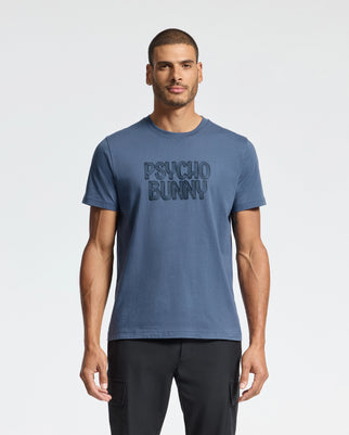 A man wearing the MENS MONARC WORDMARK GRAPHIC TEE - B6U210E200 in soft Pima cotton from Psycho Bunny, featuring "Psycho Bunny" printed on the front, stands against a plain white background. He maintains a neutral expression, paired with black pants.