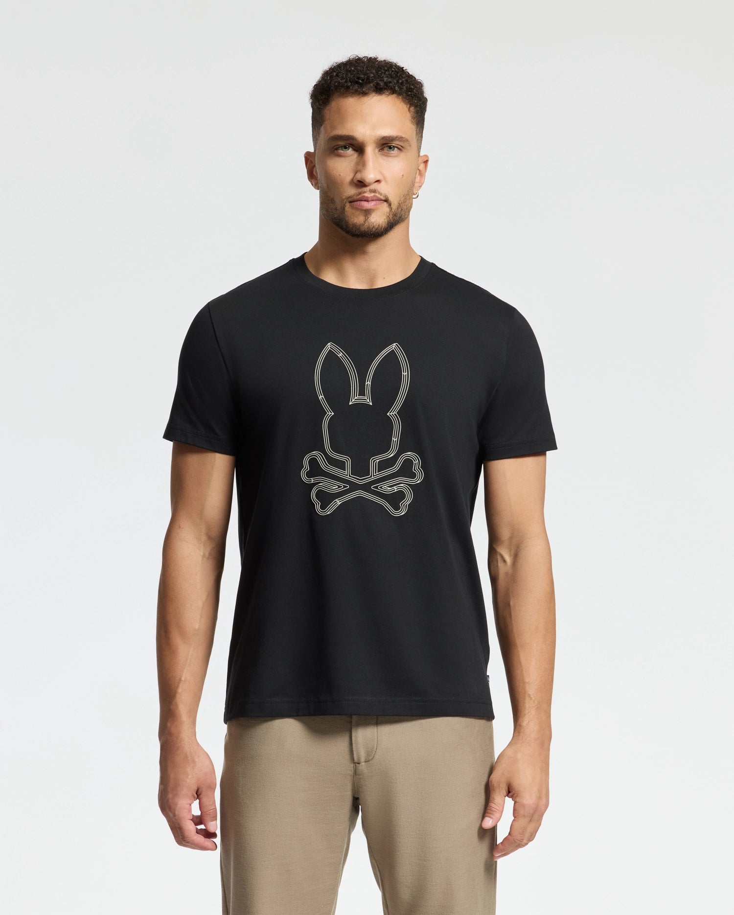 A man stands against a plain background wearing the Psycho Bunny MENS MONARC FRONT BUNNY GRAPHIC TEE - B6U211E200, made from 100% Pima cotton with a rib knit crewneck and crossbones design on the front. He pairs it with light brown pants.