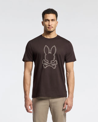 A man stands against a plain background, wearing the Psycho Bunny Men's Monarc Front Bunny Graphic Tee (B6U211E200), crafted from soft Pima cotton. The shirt showcases a raised-printed Bunny and crossbones design. His short curly hair complements his light-colored pants.