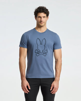 A man with curly hair stands against a plain background wearing the Psycho Bunny MENS MONARC FRONT BUNNY GRAPHIC TEE - B6U211E200, crafted from soft Pima cotton. His blue T-shirt showcases a stylized bunny and crossbones graphic and he pairs it with black pants.