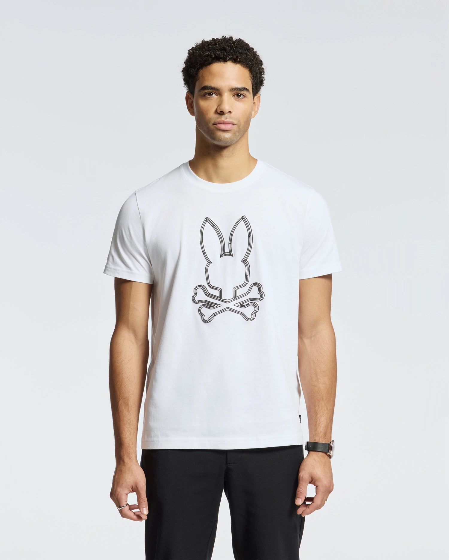 A man wears the Psycho Bunny Men's Monarc Front Bunny Graphic Tee (B6U211E200), crafted from 100% Pima cotton and made in Peru. The white T-shirt, showcasing a bunny above crossed bones design, complements his black pants and wristwatch as he stands against a plain gray background.