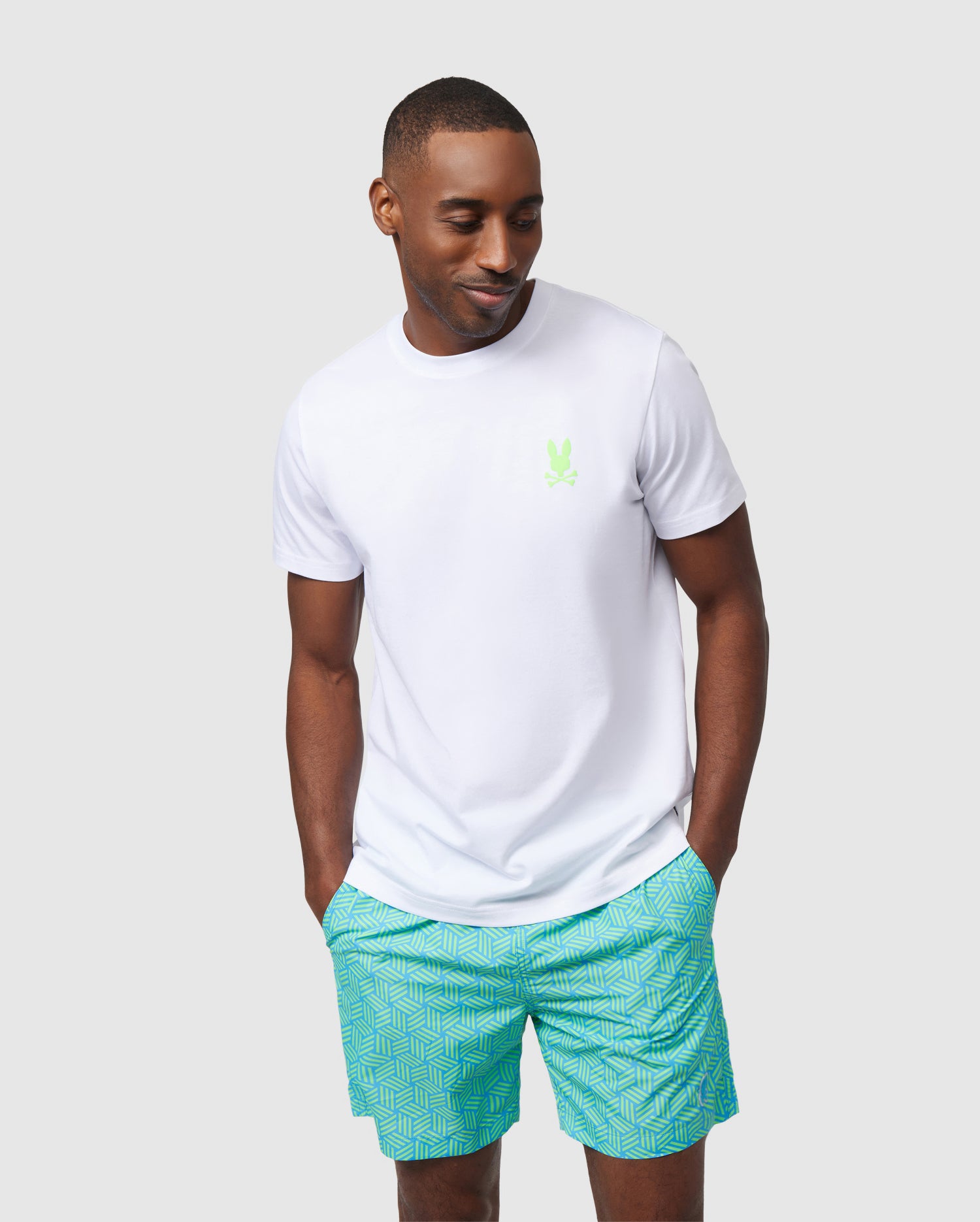 A man in a Psycho Bunny MENS SLOAN BACK GRAPHIC TEE with a small green logo and turquoise patterned shorts stands against a light gray background, looking to his right with a relaxed pose.