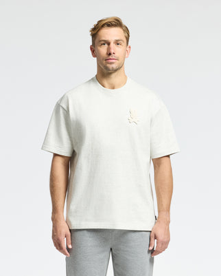 A man with short light brown hair wears a Psycho Bunny MENS ELDORADO HEAVY WEIGHT RELAXED FIT TEE in light gray, crafted from soft Peruvian Pima cotton and featuring an embroidered chest design, paired with matching gray pants against a plain white background.