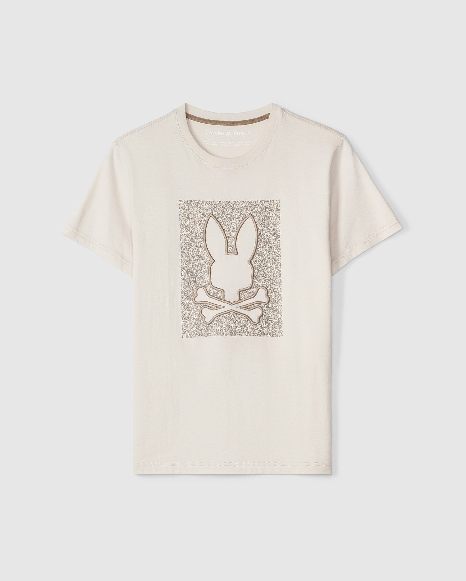 A plain light beige Psycho Bunny Livingston tee featuring a centered embroidered Bunny design of a stylized white rabbit head over crossbones, all set within a textured silver square background.