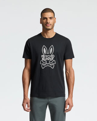 A man stands against a plain background wearing the Psycho Bunny MENS DUMON GRAPHIC TEE - B6U271E200, featuring a bunny skull and crossbones design. He complements this shirt with gray pants while maintaining a neutral expression.