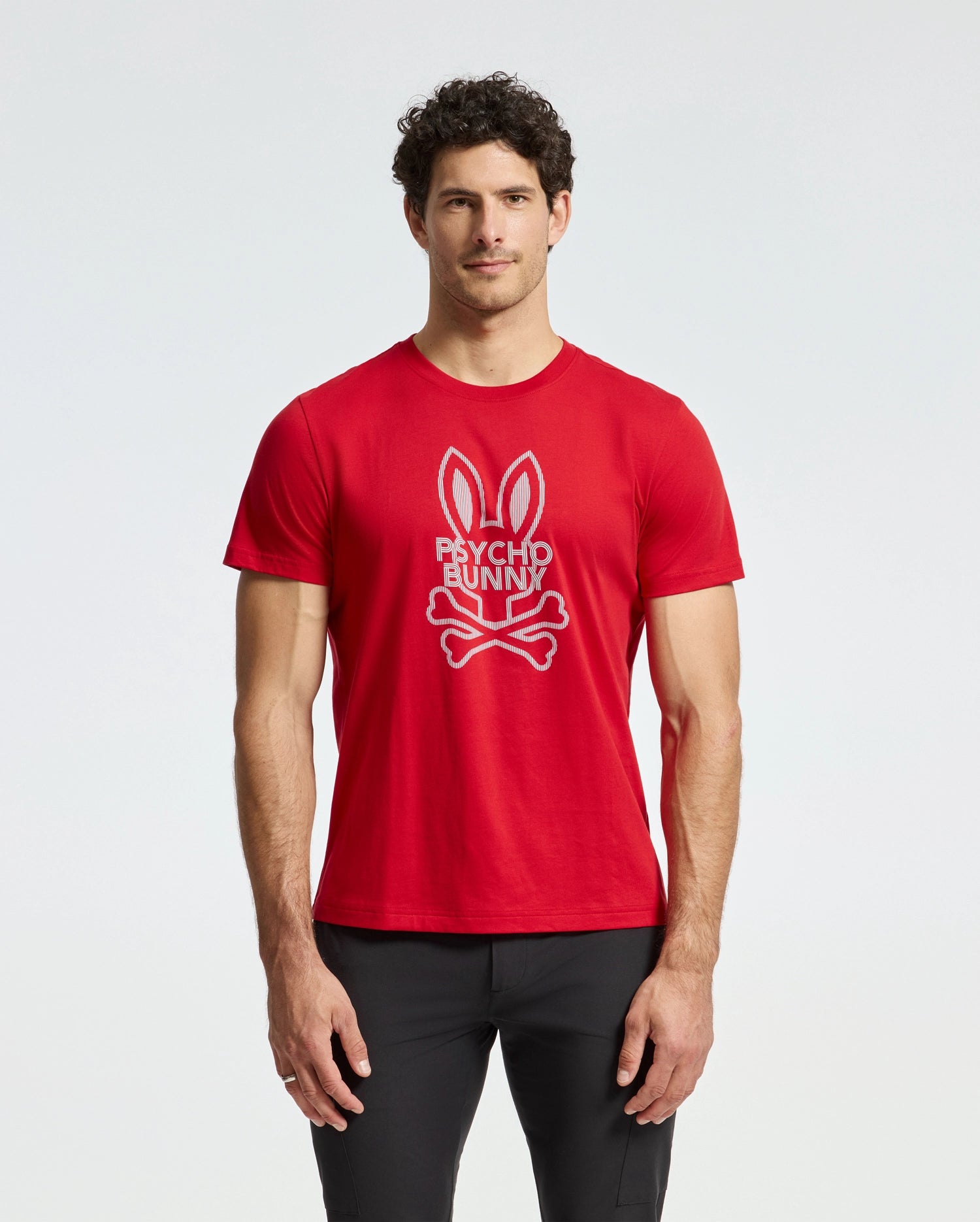 A man wearing a red MENS DUMON GRAPHIC TEE - B6U271E200 from Psycho Bunny, featuring a bunny and crossbones logo and the text 