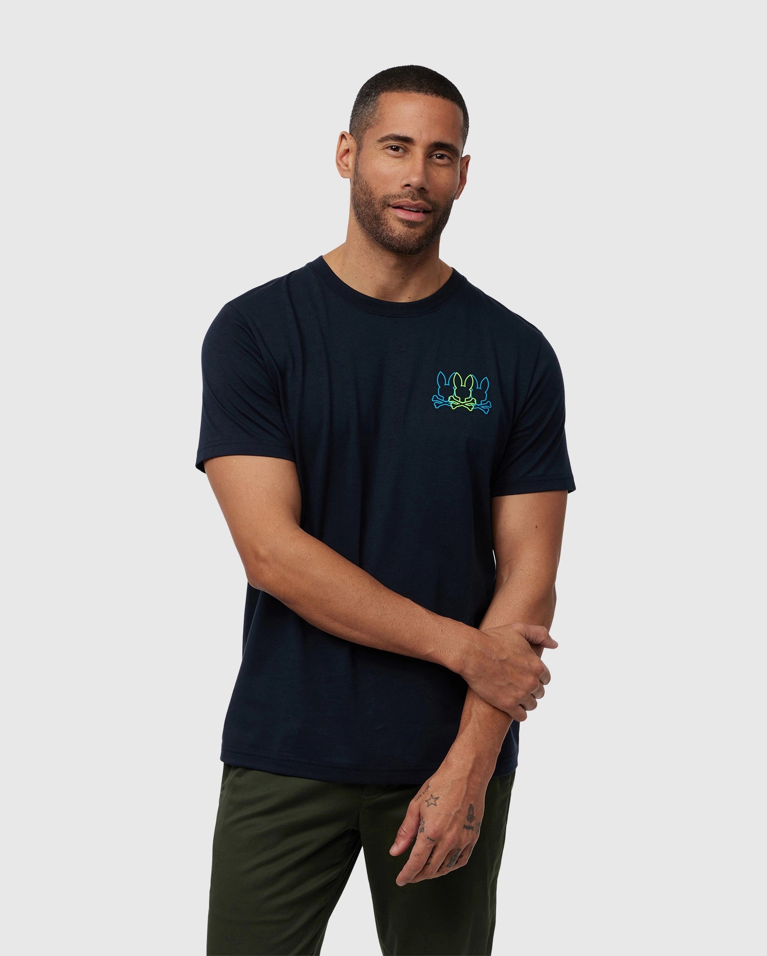 A man in a navy blue Psycho Bunny Pompey graphic tee with a small green emblem on the chest stands with arms crossed, wearing olive pants, on a plain background.