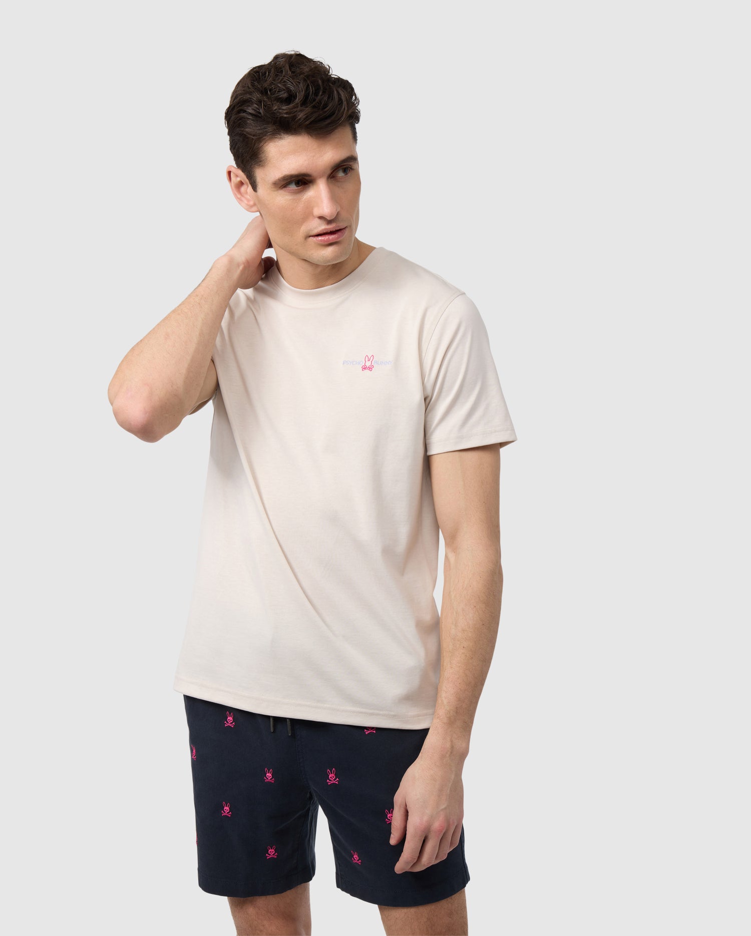 A man in a beige MENS WASTERLO BACK GRAPHIC TEE from Psycho Bunny with an embroidered chest logo and black shorts with pink floral embroidery stands against a white background, looking to the side and touching the back of his neck.