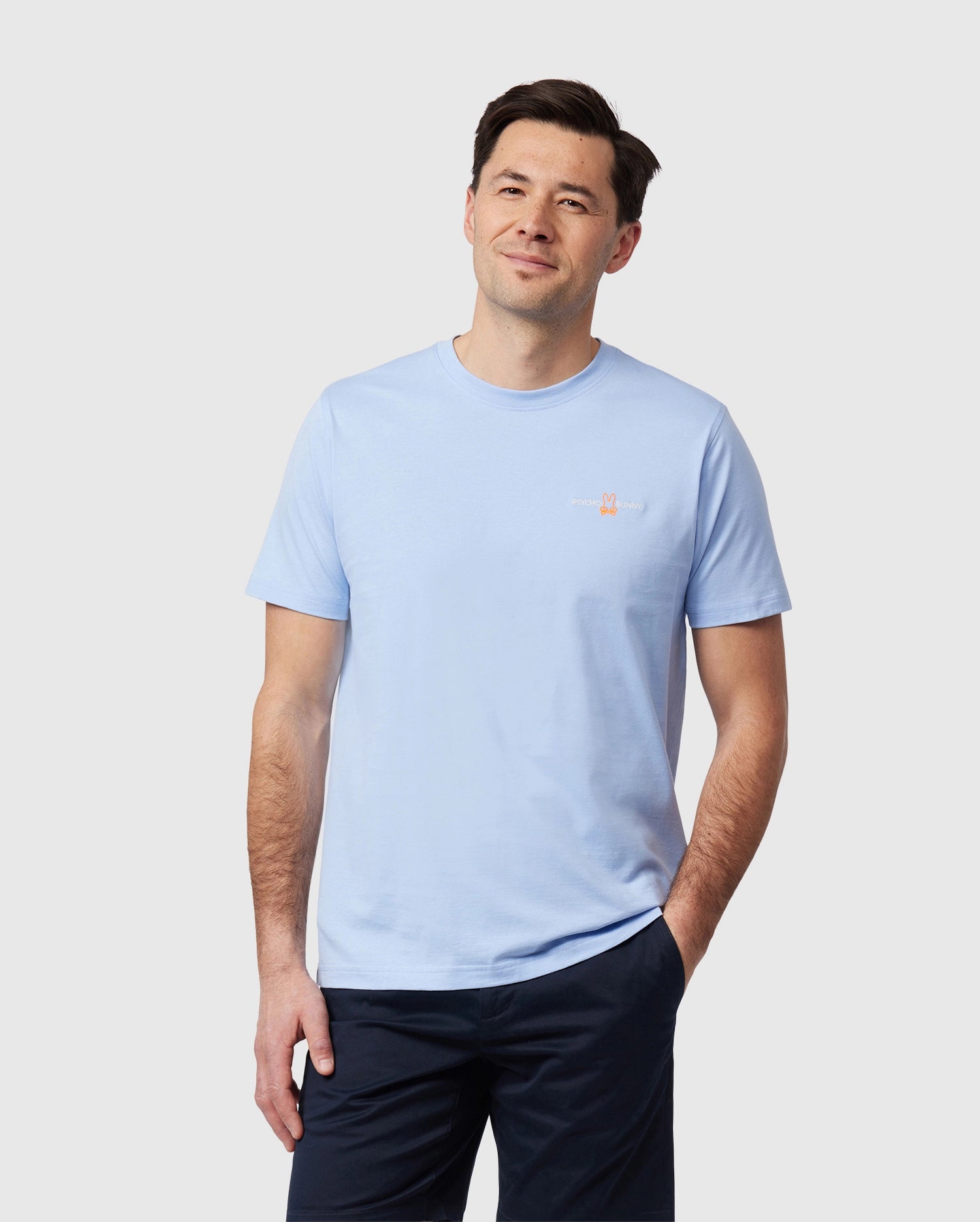 A man with short dark hair is standing against a plain background, wearing a light blue Psycho Bunny MENS WASTERLO BACK GRAPHIC TEE - B6U317B2TS made from Peruvian Pima cotton and dark pants. He has one hand in his pocket and is smiling gently, looking straight ahead.