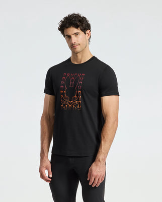 A man with curly hair is wearing a black MENS CORTES GRAPHIC TEE - B6U328E200 by Psycho Bunny, showcasing a vibrant design on the front. He stands against a simple, light gray background, gazing toward the camera.