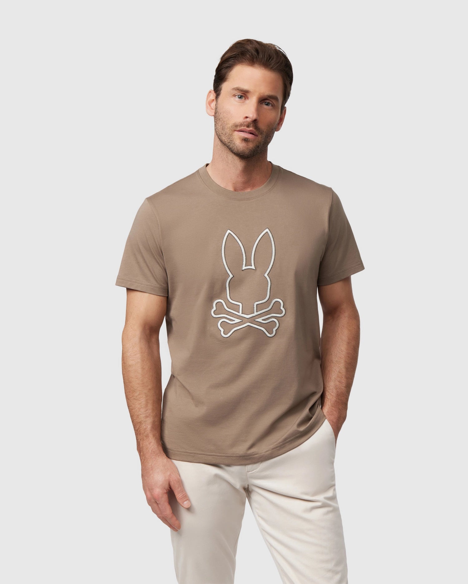 A man with a beard modeling a brown Psycho Bunny graphic tee (MENS FLOYD GRAPHIC TEE - B6U338B2TS) with an embroidered bunny skull and crossbones logo, paired with light khaki pants, standing against a plain background.