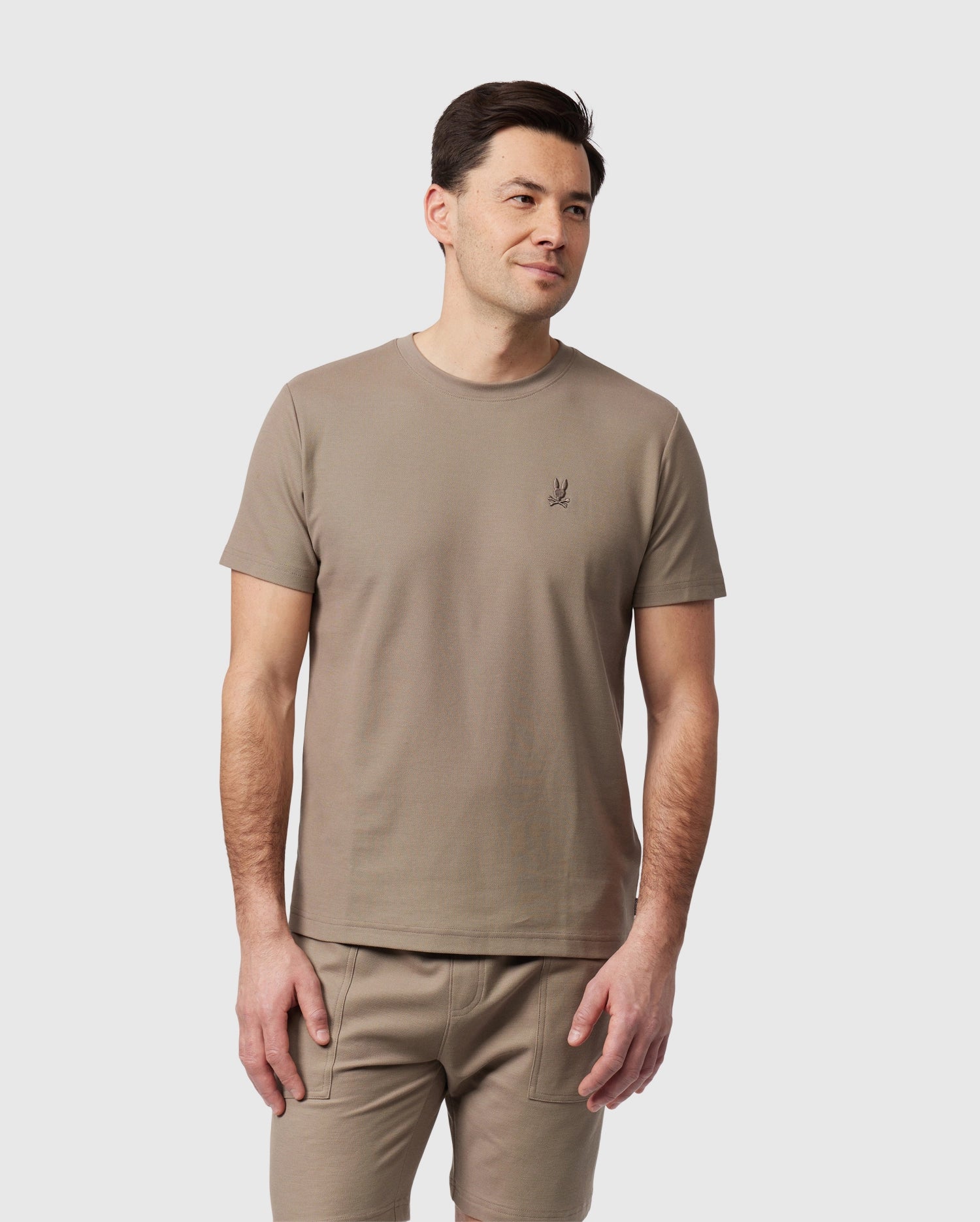 A man with short dark hair is standing against a plain white background. He is wearing a beige Men's Stanford Pique Tee - B6U340B200 by Psycho Bunny made of diamond-knit Pima cotton, featuring a small, dark plant logo on the left side of his chest, and matching beige pants. He has a neutral expression and his arms are relaxed by his sides.