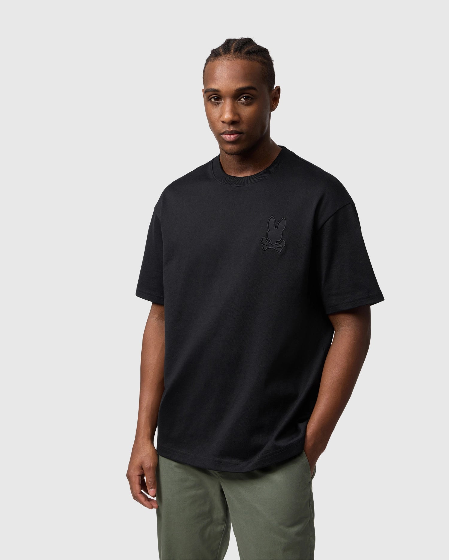 A man wearing a Psycho Bunny MENS DANBY OVERSIZED TEE - B6U359B2TS with a small embroidered logo and olive green pants stands against a light gray background, looking at the camera with a neutral expression.